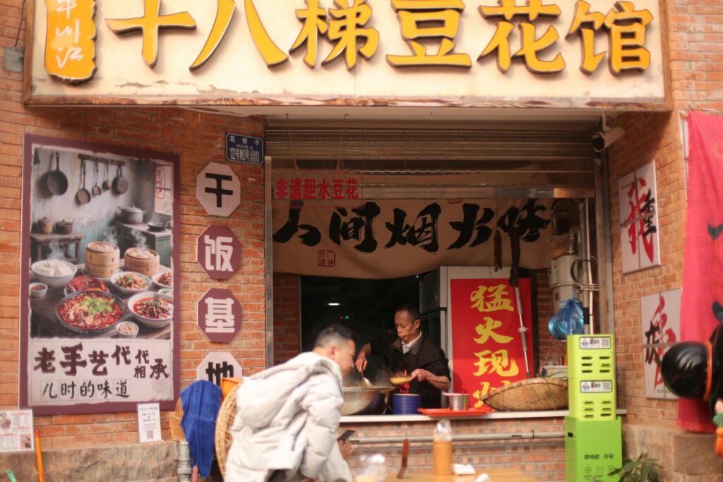 A chef prepares steaming food inside a brick-fronted eatery adorned with large yellow signage and posters of traditional dishes. Passersby walk in the foreground while various banners and menus.