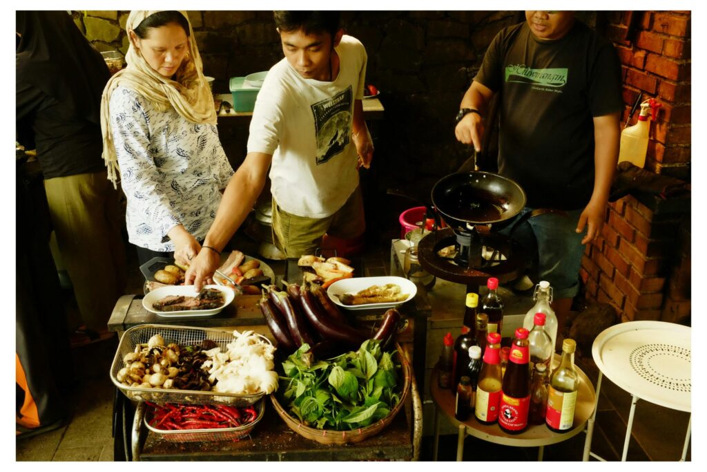 A group of people are gathered in an outdoor kitchen area, busy preparing various dishes with fresh ingredients like eggplant, mushrooms, and peppers. One man stirs a wok over a portable stove while others arrange plates of cooked food and select sauces.