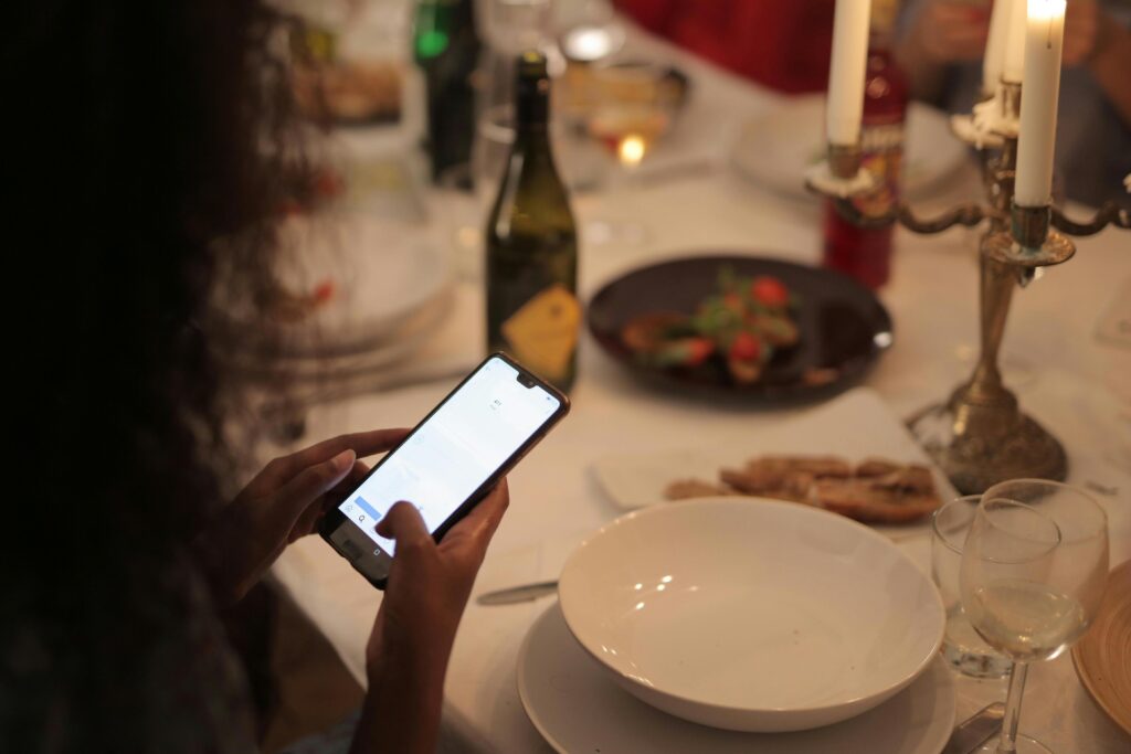 A person sits at a dinner table and checks their smartphone amidst a spread of food, wine, and lit candles. The background is softly blurred, focusing on the glowing screen and the elegant table setting.