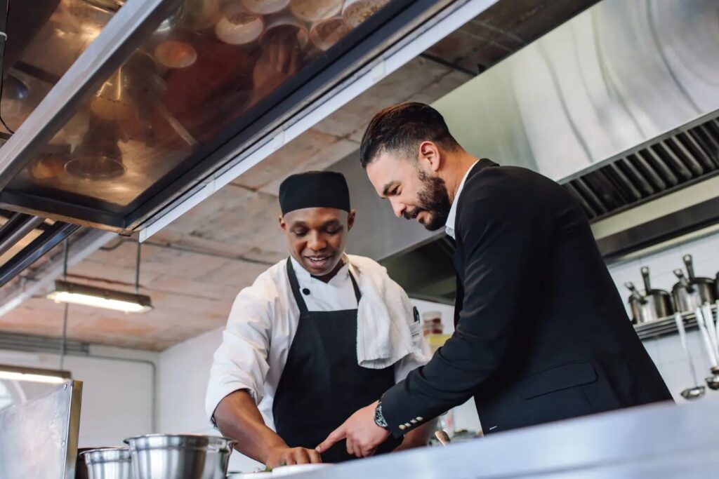 A man in a professional suit stands in a commercial kitchen, pointing toward a dish as he talks with a chef. The chef, wearing a black apron and cap, looks down at the counter while listening to the man's feedback.