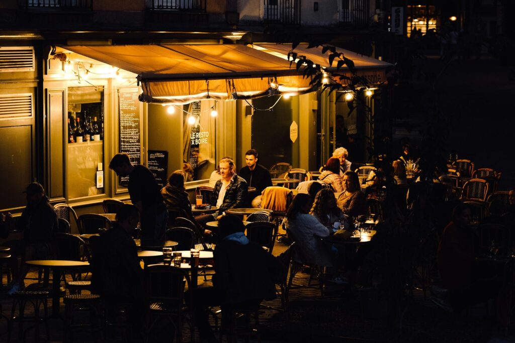 A crowd of people gathers at an outdoor cafe at night, illuminated by the warm, golden glow of string lights and an awning. Patrons sit at small round tables, creating a cozy and bustling atmosphere.