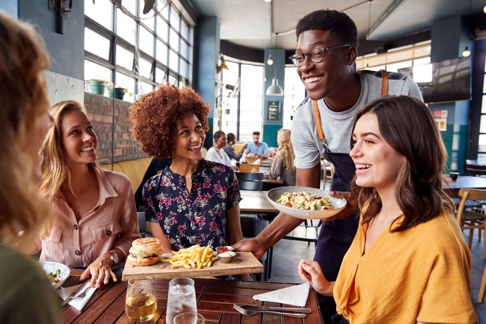 A smiling waiter serves a salad and a burger with fries to a group of four women at a bright, sunlit restaurant table. The atmosphere is warm and social, capturing a candid moment of laughter and friendly interaction
