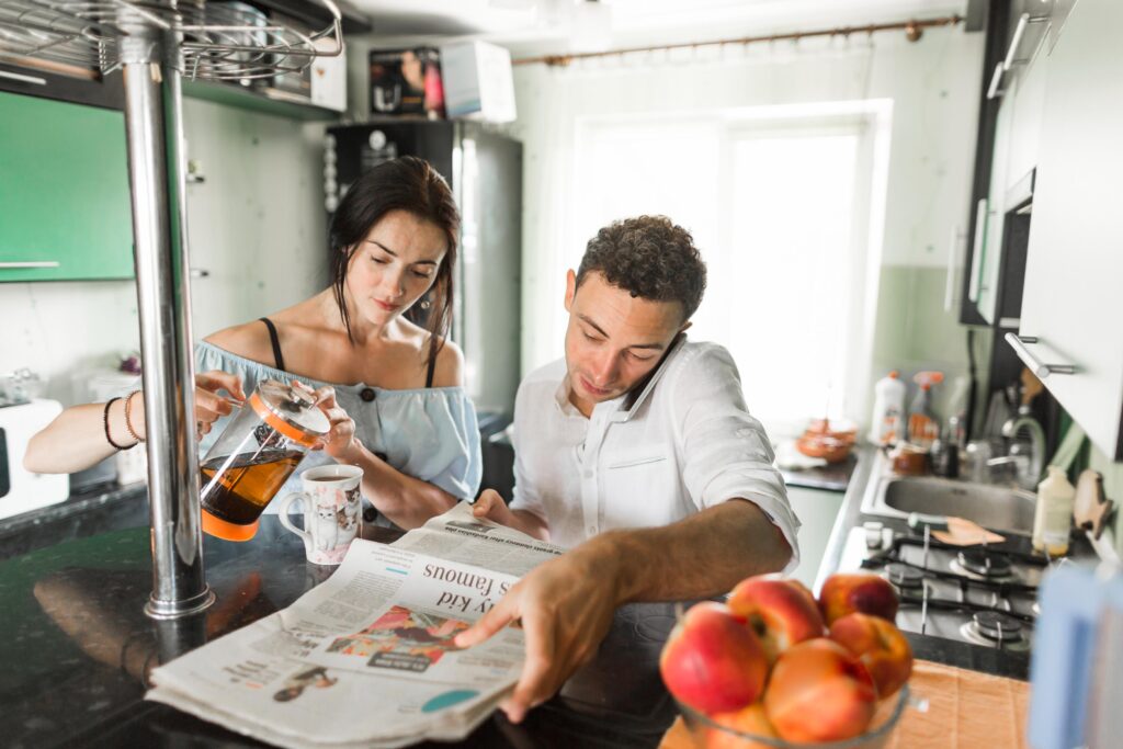 A woman pours tea from a French press into a cat-themed mug while her partner talks on a mobile phone. The couple is gathered in a bright kitchen, with a newspaper spread out on the counter and a bowl of fresh peaches in the foreground.