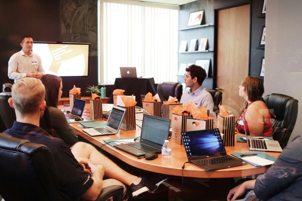 A group of professionals sits around a conference table with laptops and gift bags while a man presents information on a screen at the front of the room.
