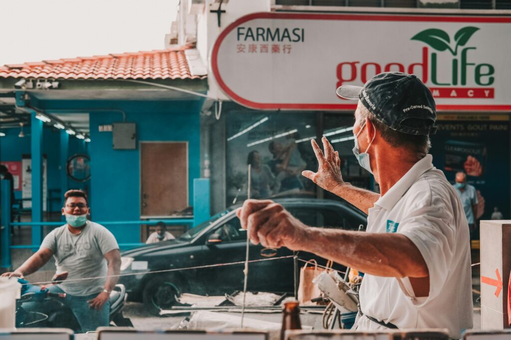 A man in a face mask and cap works at an outdoor food stall, gesturing with flour-dusted hands. In the background, a person on a motorbike passes by a pharmacy