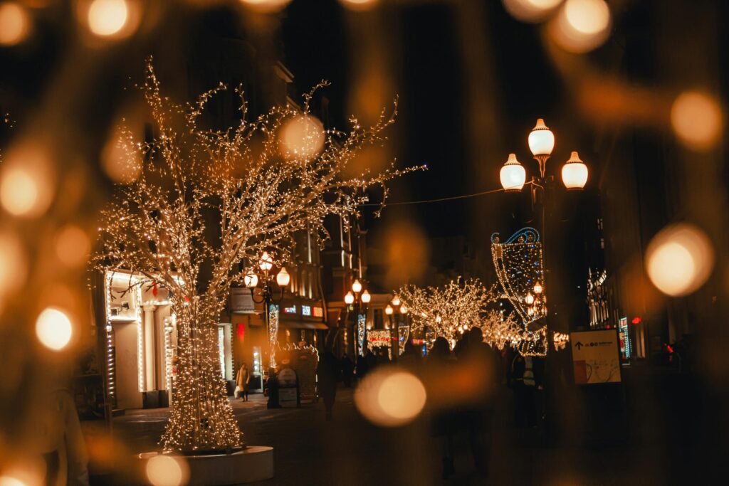A vibrant city street is transformed into a winter wonderland with trees draped in shimmering golden fairy lights. Pedestrians stroll through the warm, festive glow as soft bokeh orbs frame the enchanting nighttime scene.