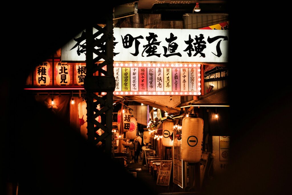 A vibrant collection of glowing Japanese lanterns and illuminated shop signs creates a bustling night scene tucked beneath a dark structural frame. The warm lights and traditional calligraphy invite viewers into a narrow, lively marketplace filled with texture and character.