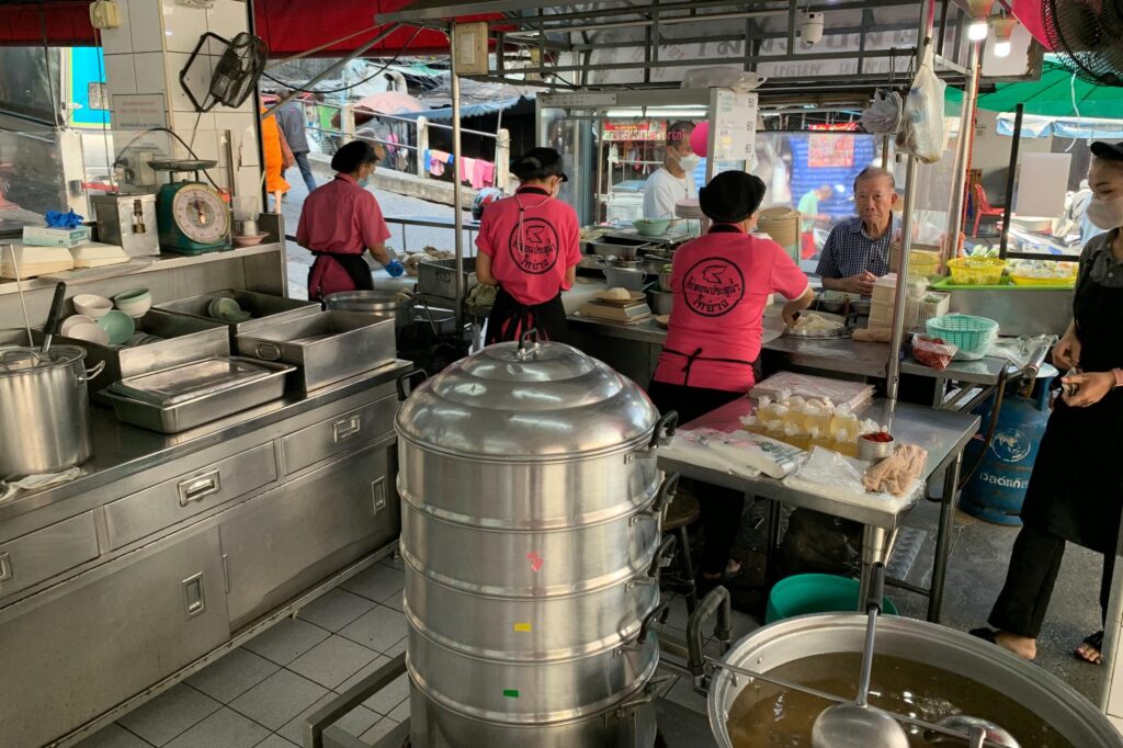 Staff members in matching pink uniforms prepare food at a busy open-air street stall featuring large metal steamers and stainless-steel workstations. In the background, an older man sits at a small table while pedestrians and street activity are visible