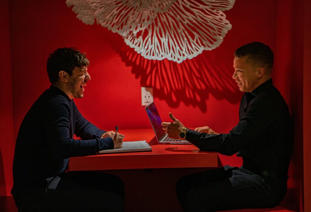 Two men sit across from each other at a vibrant red table in a modern, dimly lit space, seemingly engaged in a lively business discussion or interview. One man laughs while taking notes in a notebook, while the other gestures toward an open laptop.