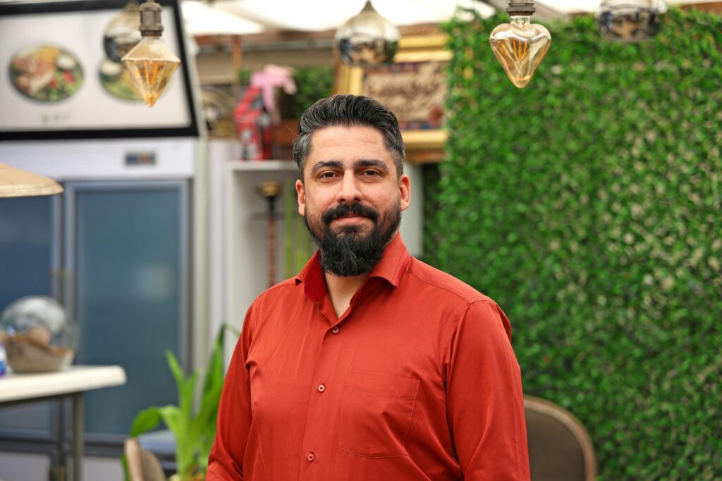 A man with a dark beard and a red button-down shirt stands in what appears to be a restaurant or cafe setting.