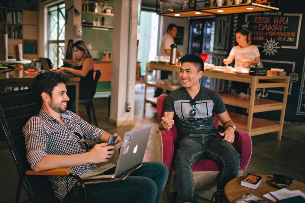 Two men sit in a bustling, modern cafe, smiling and engaging in conversation while using a laptop. In the background, other patrons work and staff members prepare drinks near a chalkboard menu.