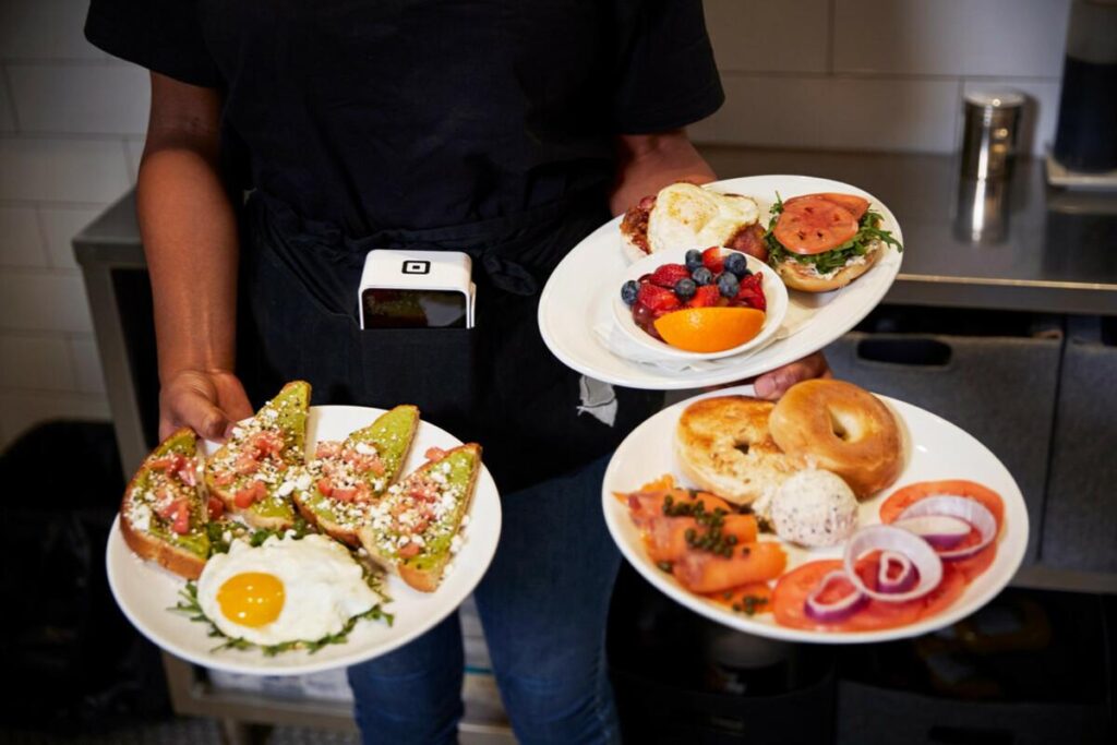 A person in a black apron is skillfully holding three plates filled with various breakfast dishes, including avocado toast, bagels with lox, and fresh fruit. A white mobile card reader is tucked into their apron pocket, suggesting a busy restaurant or cafe environment.