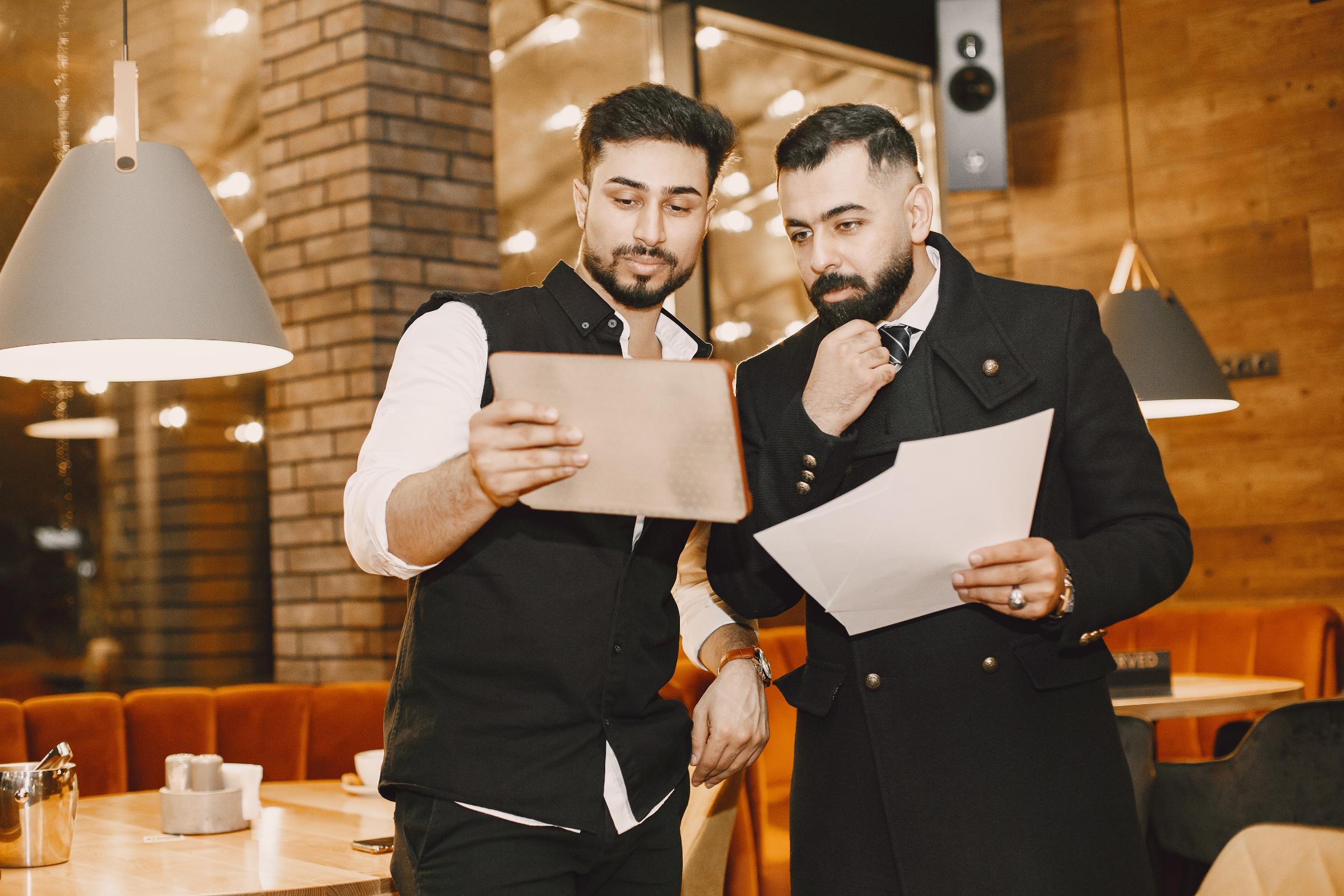 Two men are standing in a restaurant while looking intently at a tablet and some papers. They appear focused on their discussion amidst the warm, modern interior of the establishment.