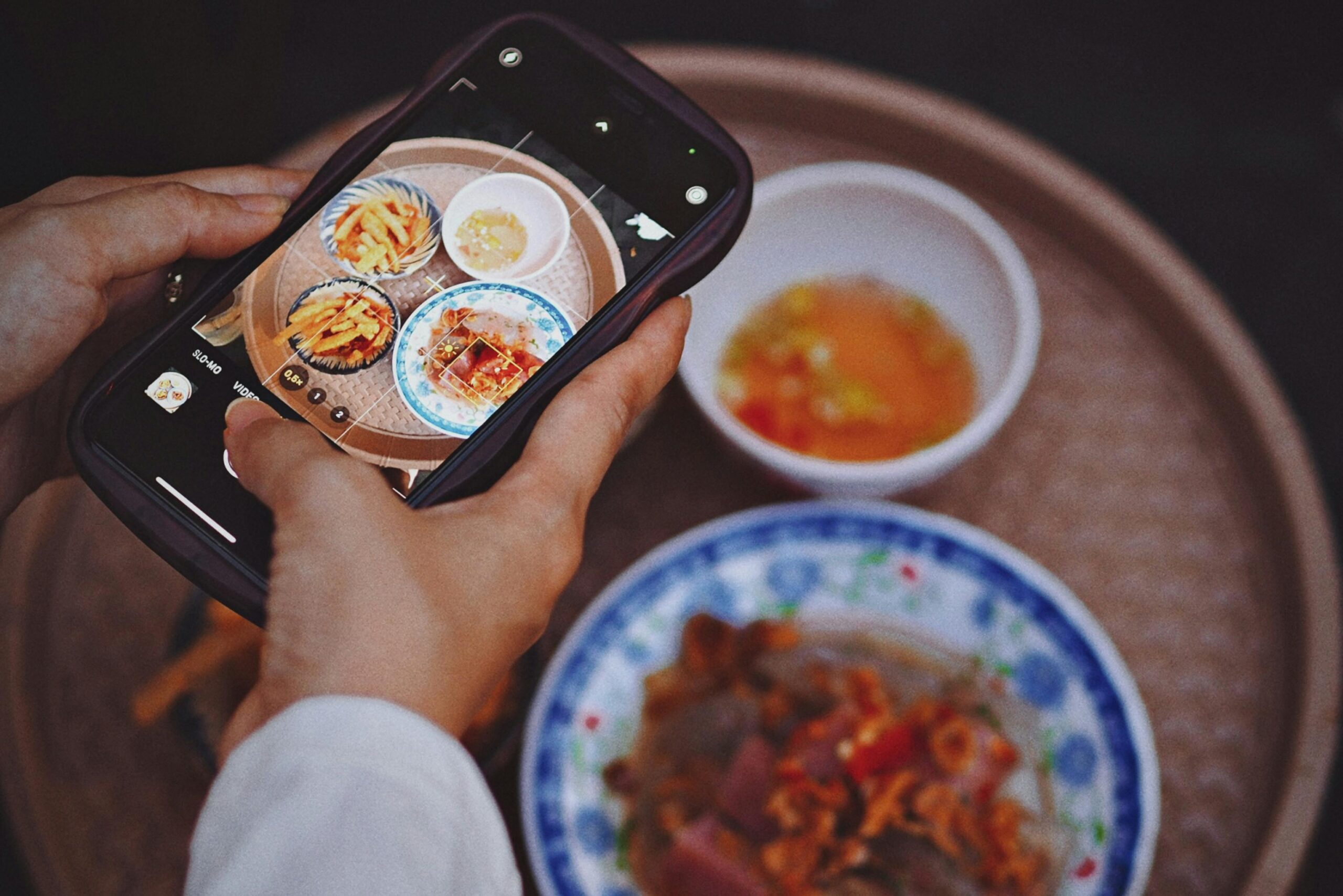 A person holds a smartphone over a tray to capture a top-down photo of several Vietnamese dishes, including small bowls of dipping sauce and savory snacks. The image features a shallow depth of field.