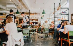 This brightly lit cafe features an industrial aesthetic with exposed piping, sage green chairs, and shelves stocked with various retail goods. Patrons are scattered throughout the space.
