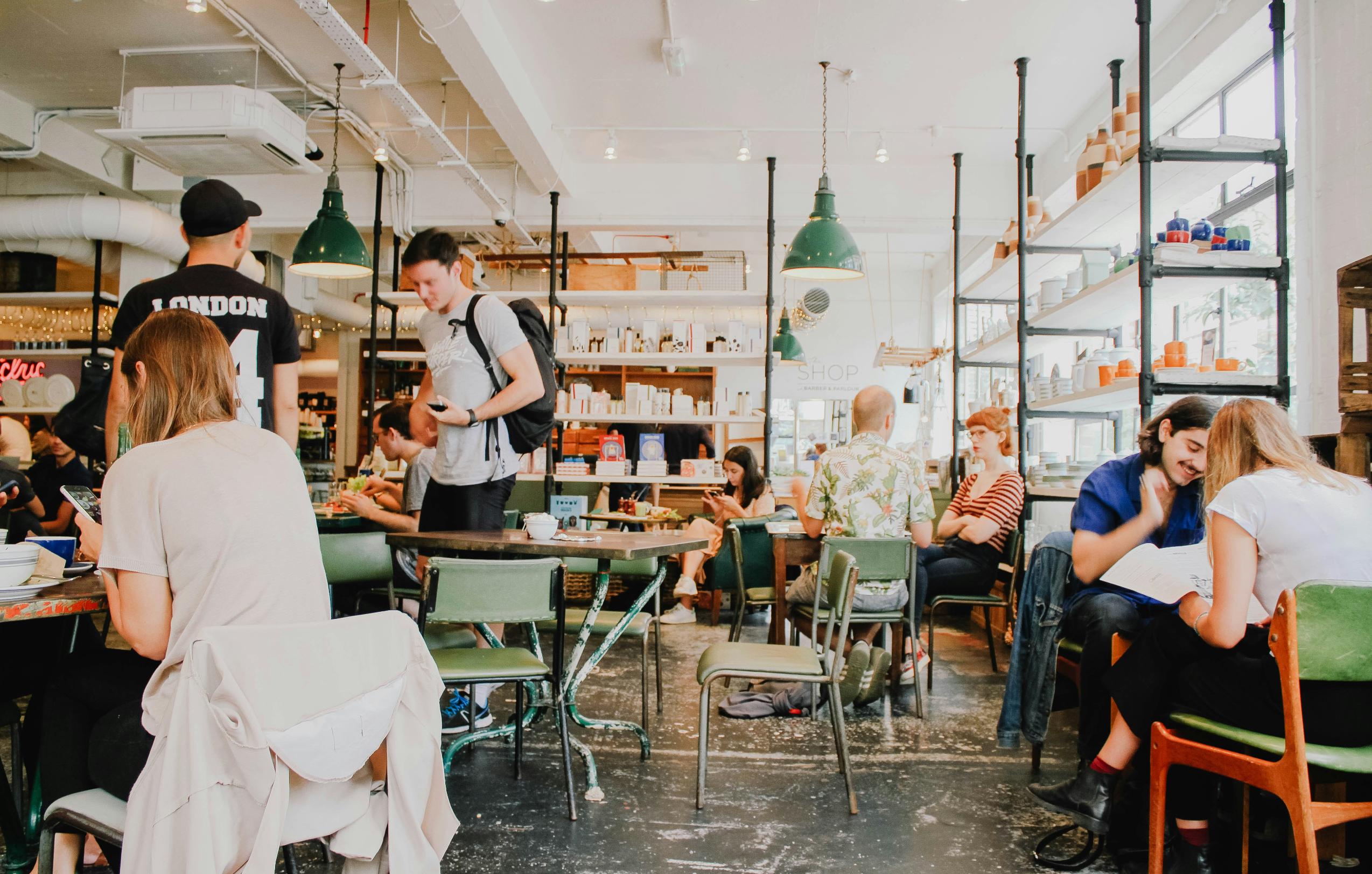 This brightly lit cafe features an industrial aesthetic with exposed piping, sage green chairs, and shelves stocked with various retail goods. Patrons are scattered throughout the space.