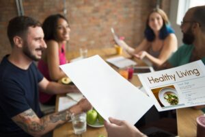 A group of people are gathered around a wooden table in a brightly lit room, engaged in a discussion while holding papers focused on healthy living.
