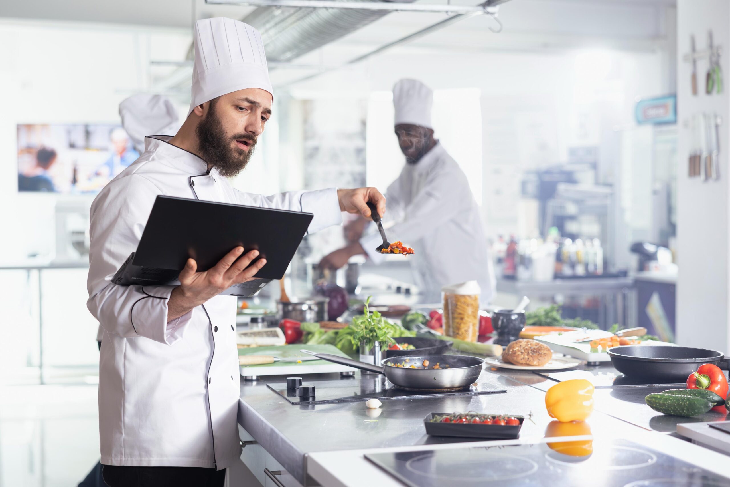In a busy professional kitchen, a bearded chef in a white uniform holds a laptop while carefully tasting or inspecting food from a small spoon. Surrounded by fresh vegetables and stainless steel counters.