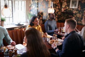 A group of friends enjoys a meal and conversation while seated in a cozy, booth-lined restaurant featuring floral wallpaper. Plates of classic pub-style food, such as savory pies and roasted chicken, are spread across the wooden tables under warm pendant lighting.