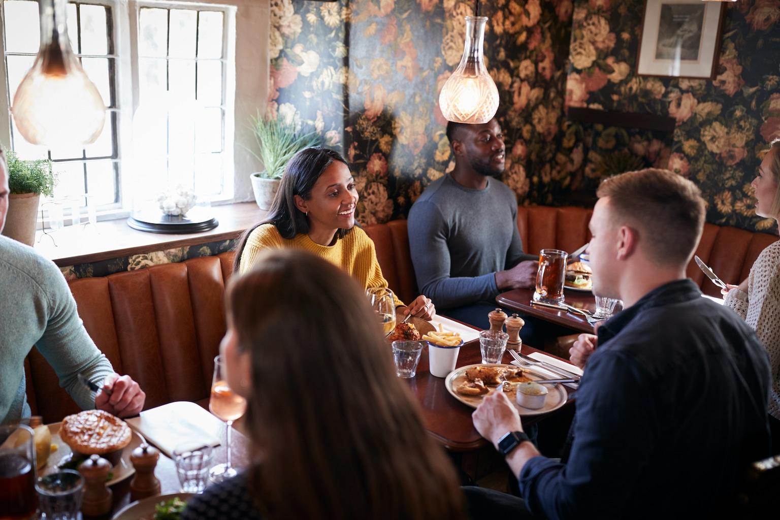 A group of friends enjoys a meal and conversation while seated in a cozy, booth-lined restaurant featuring floral wallpaper. Plates of classic pub-style food, such as savory pies and roasted chicken, are spread across the wooden tables under warm pendant lighting.
