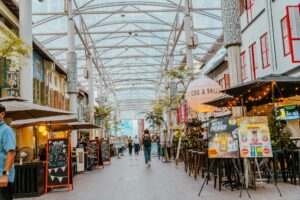 A covered pedestrian walkway is lined with outdoor restaurants, promotional signs, and white pillars under a glass roof. People walk through the open-air space, which features a mix of modern architecture and colorful traditional shutters.