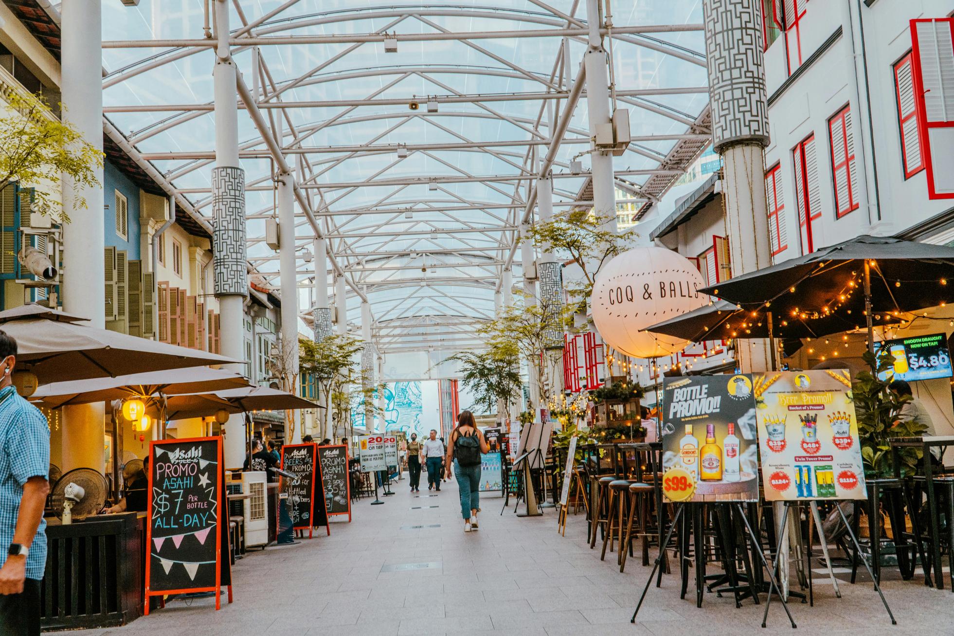 A covered pedestrian walkway is lined with outdoor restaurants, promotional signs, and white pillars under a glass roof. People walk through the open-air space, which features a mix of modern architecture and colorful traditional shutters.
