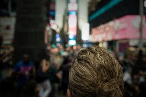 The back of a person's head with wavy blonde hair in a bun is shown in sharp detail. Behind them, a crowded city square like Times Square appears as a blur of colorful neon lights.