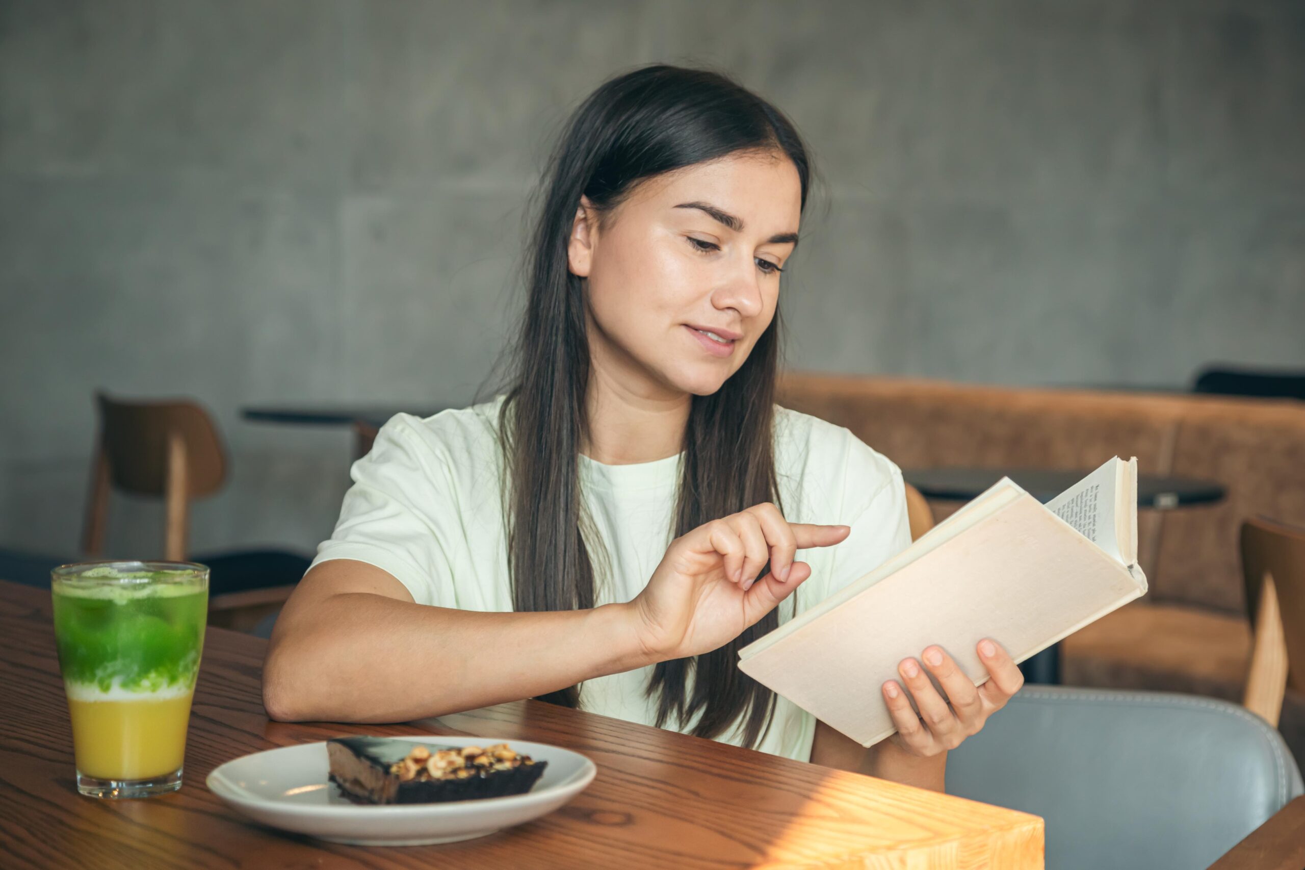 A woman with long dark hair sits at a wooden table in a cafe, focused on reading a book. A layered green and yellow drink and a slice of chocolate tart are placed on the table in front of her.