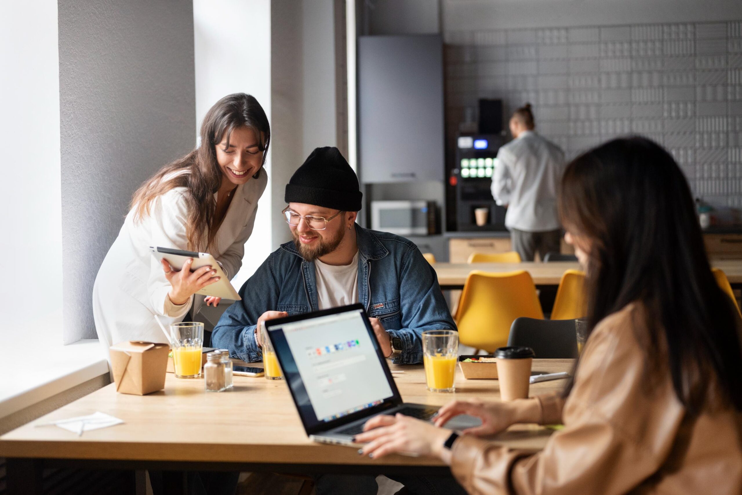 A group of colleagues enjoys a casual break in a modern office cafeteria, gathered around a wooden table with laptops, tablets, and fresh orange juice.