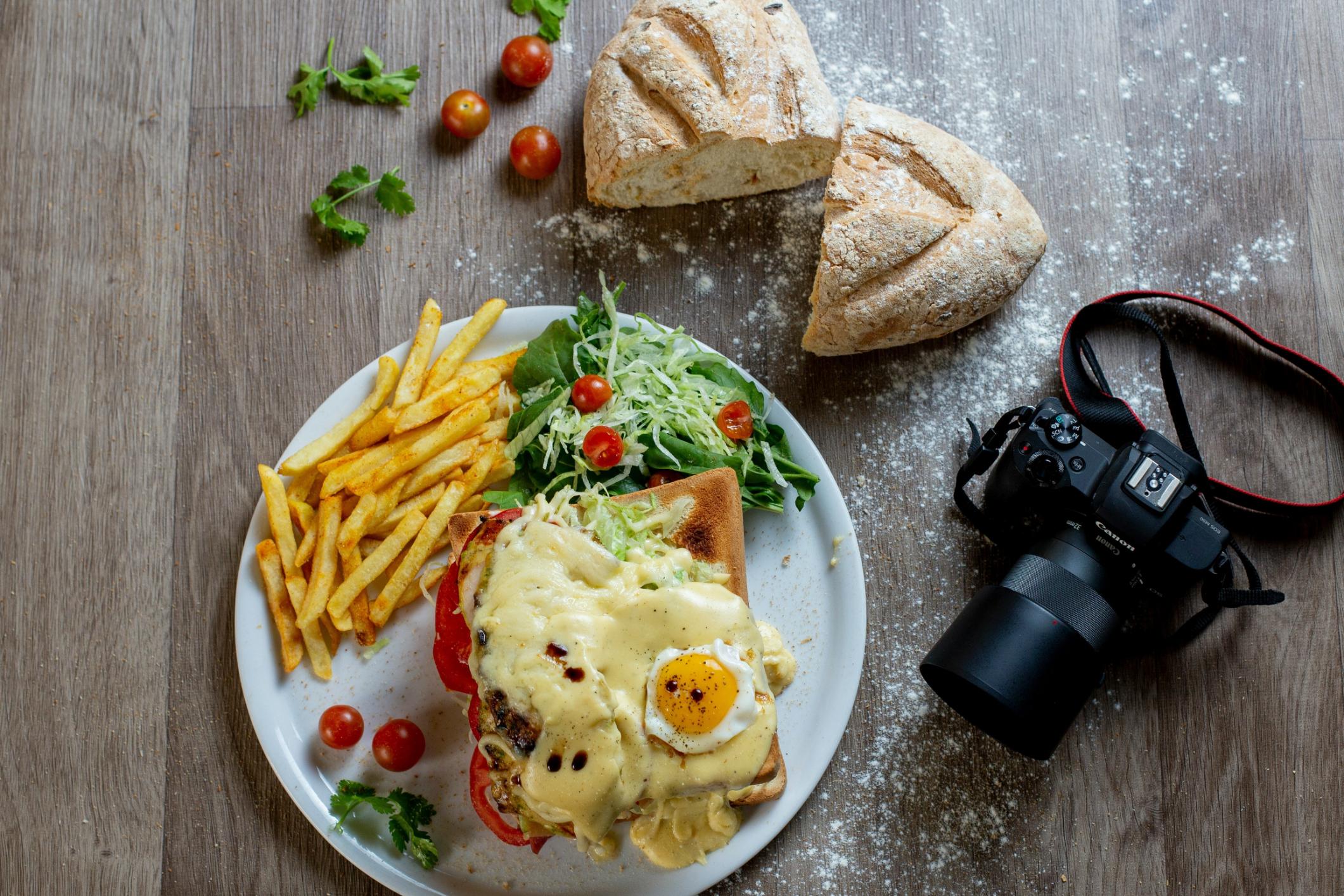 A white plate holds a cheesy open-faced sandwich topped with a fried egg, accompanied by a side of french fries and a fresh salad. Nearby on the wooden surface sits a rustic loaf of bread and a professional Canon DSLR camera.