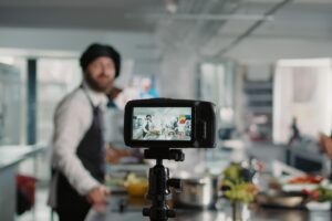 A digital camera mounted on a tripod captures a professional chef in a blurred commercial kitchen during a cooking demonstration. The camera’s display shows a wide shot of two people preparing a meal amidst various ingredients.