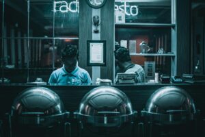 Two men work behind a counter featuring three large, silver chafing dishes. The scene is cast in a moody, deep teal light with glowing neon signs in the background.