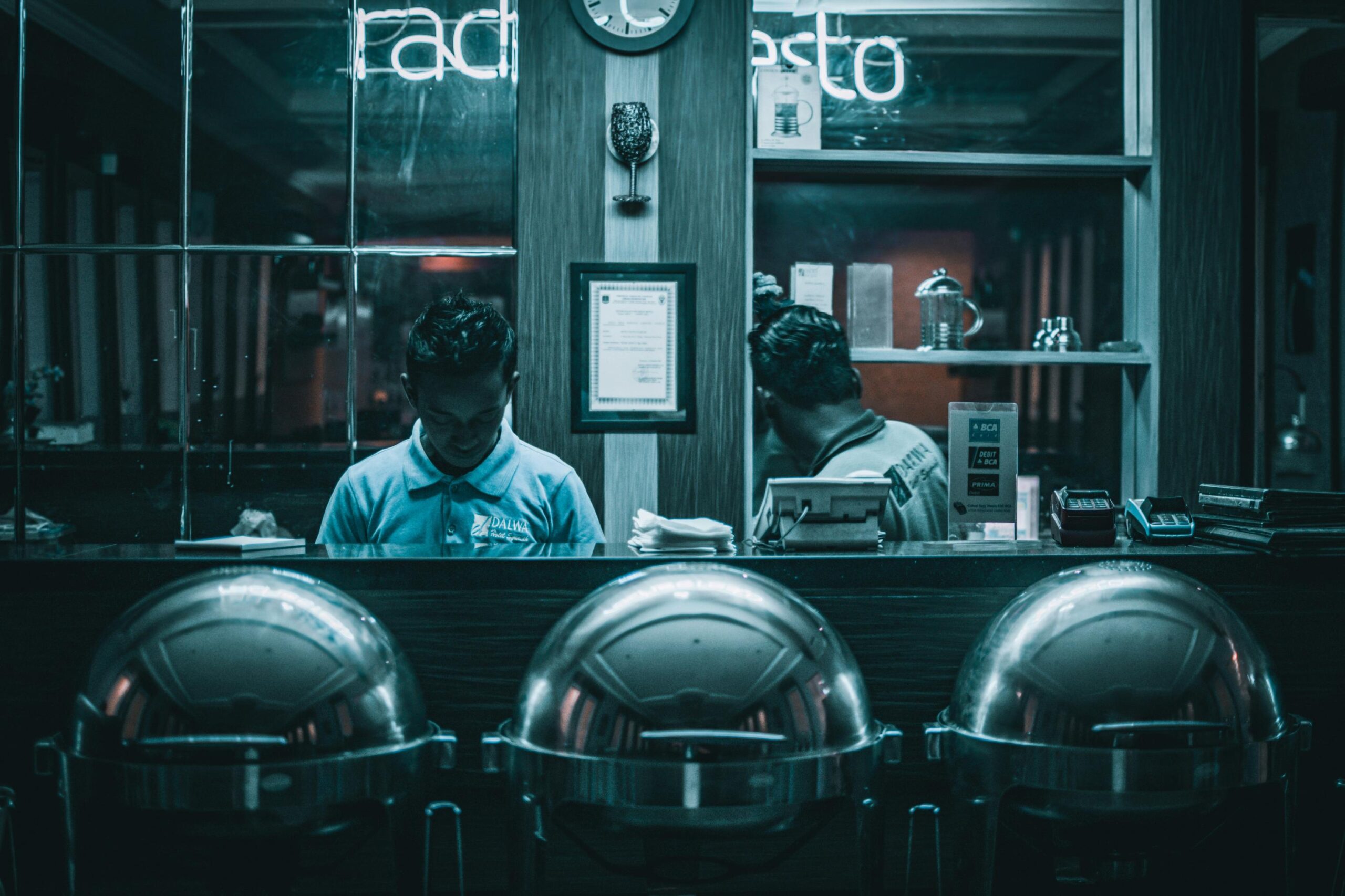 Two men work behind a counter featuring three large, silver chafing dishes. The scene is cast in a moody, deep teal light with glowing neon signs in the background.