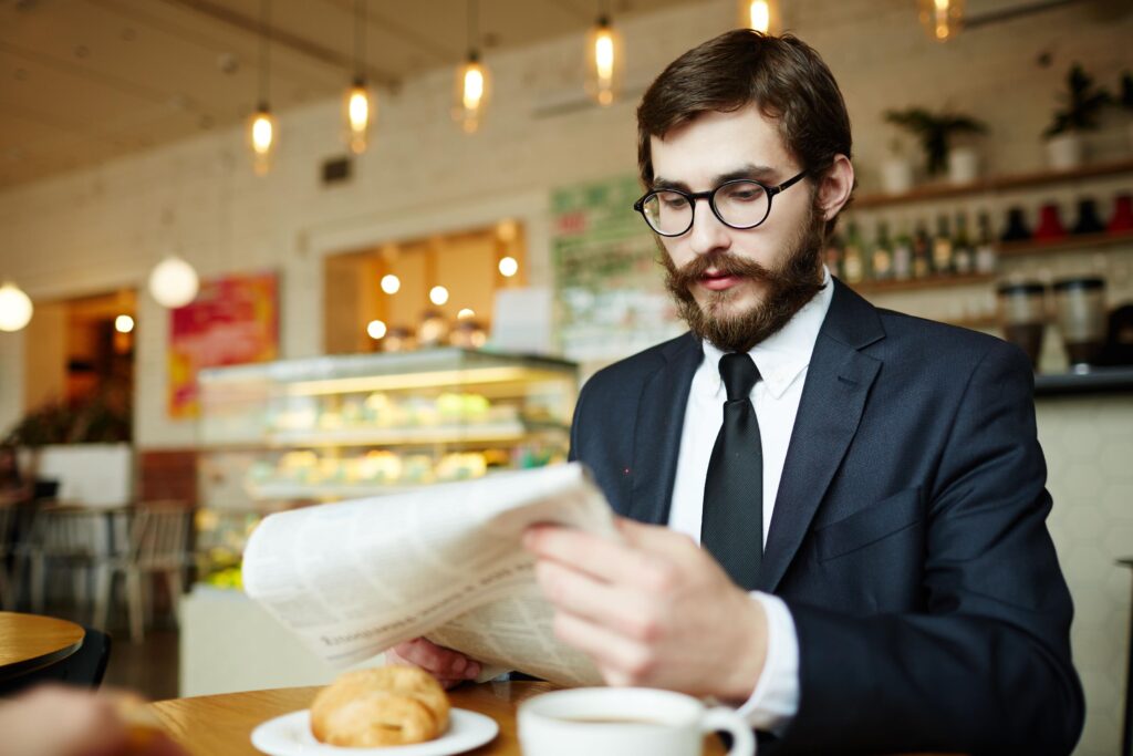A well-dressed man with a beard and glasses reads a newspaper in a café. He sits at a wooden table with a pastry and a cup of coffee.