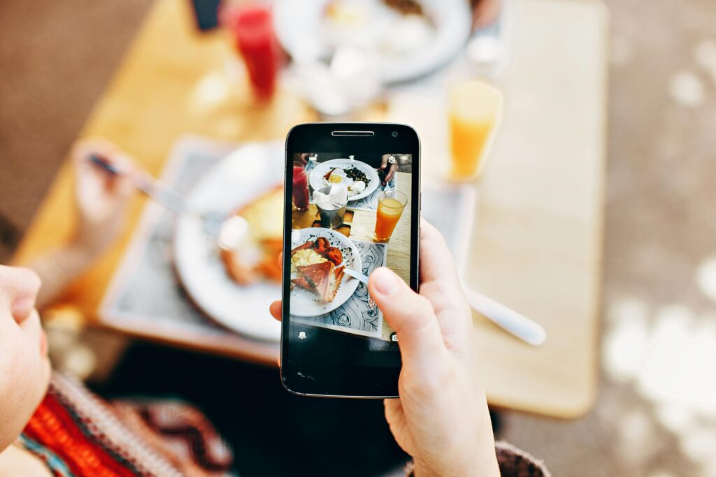 A person holds a smartphone to capture a top-down photo of a brunch spread featuring eggs, toast, and orange juice. The background shows a soft-focus view of the wooden table.