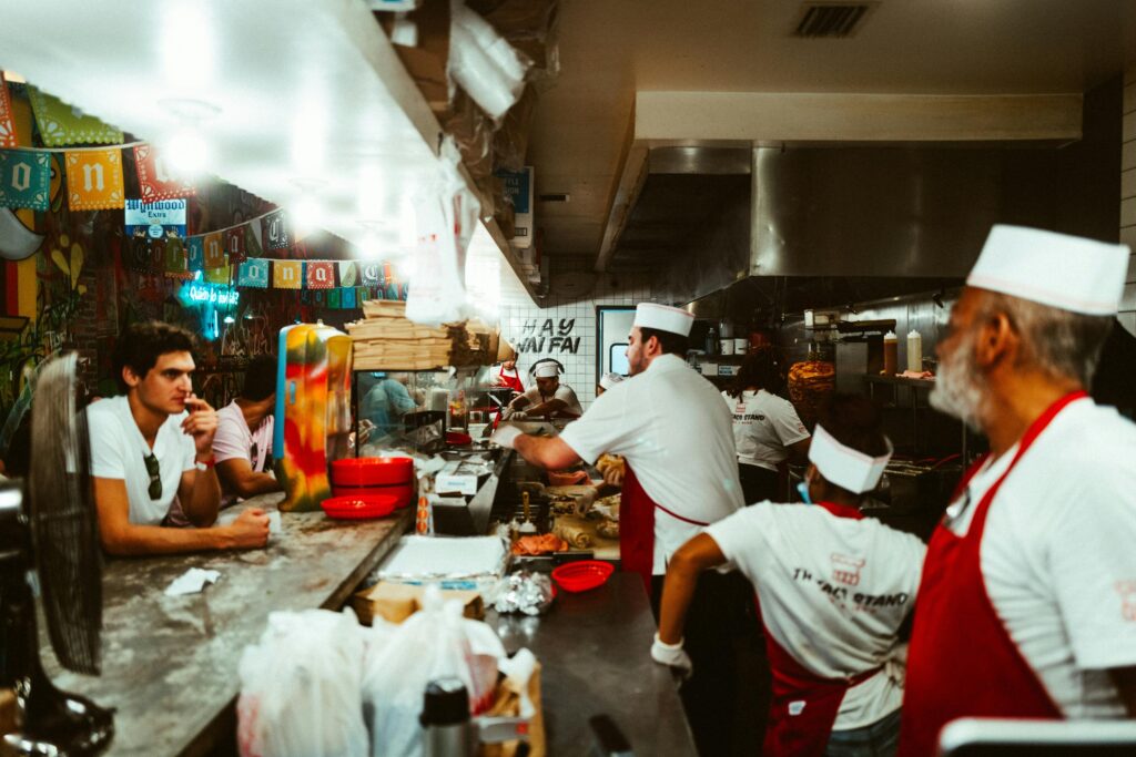 This candid shot captures the bustling atmosphere of a busy taco shop, where staff in white uniforms and red aprons prepare food behind a cluttered counter. A customer sits at the bar to the left.