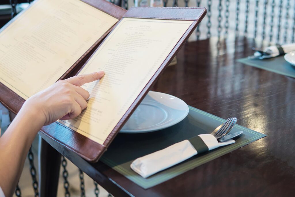 A person points a finger at a specific item on a large, leather-bound menu while seated at a dark wooden restaurant table. The table is neatly set with a white plate and silverware wrapped in a napkin on a green placemat.
