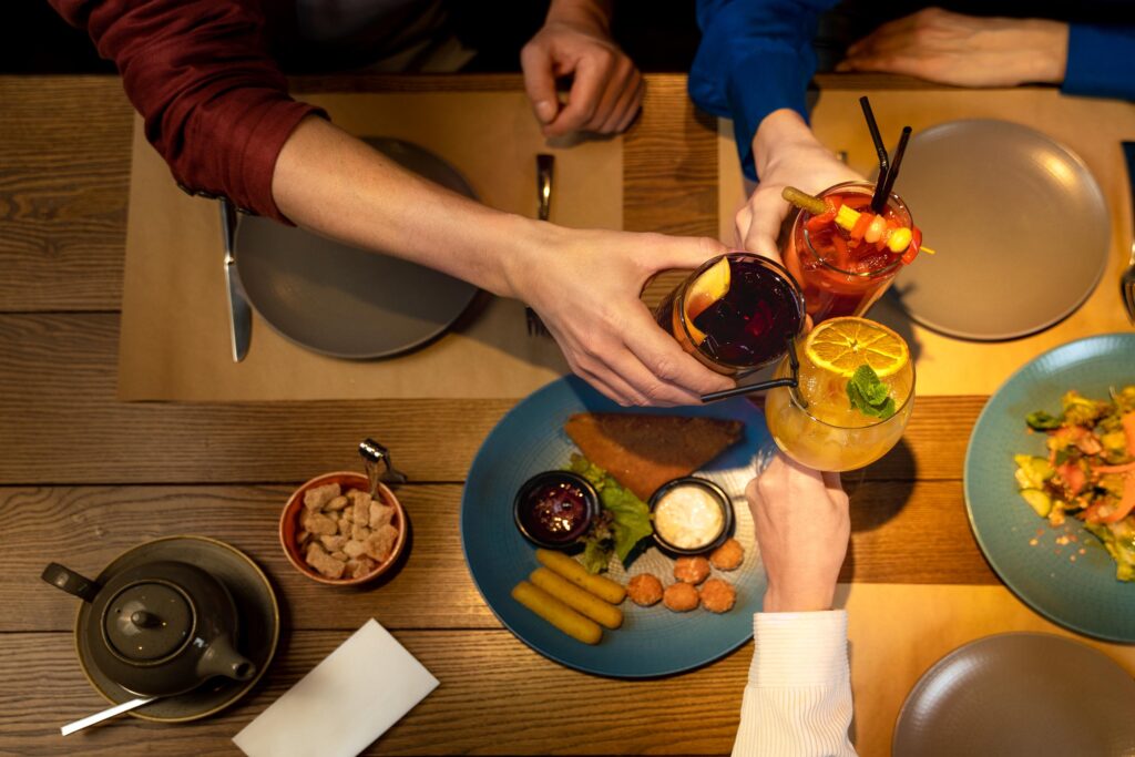 From a top-down perspective, three people are seen clinking vibrant, garnished cocktails over a wooden table spread with various appetizers and plates. The warm lighting highlights a cozy dining atmosphere.