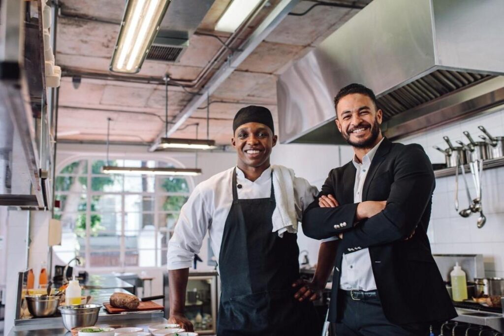 A chef in a white uniform and a man in a black suit stand side-by-side and smile in a bright, modern commercial kitchen. The industrial space features stainless steel equipment, exposed ceiling fixtures, and large windows that let in natural light.