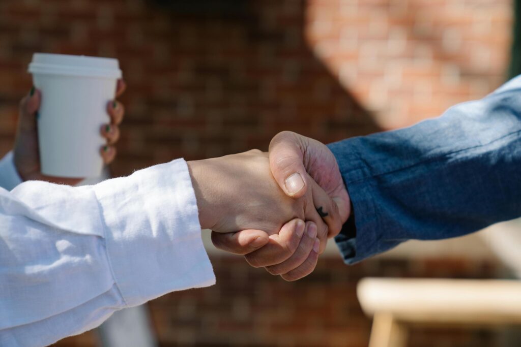 Two people shake hands in a close-up shot outdoors. One person wears a white long-sleeved shirt, while the other wears blue denim.