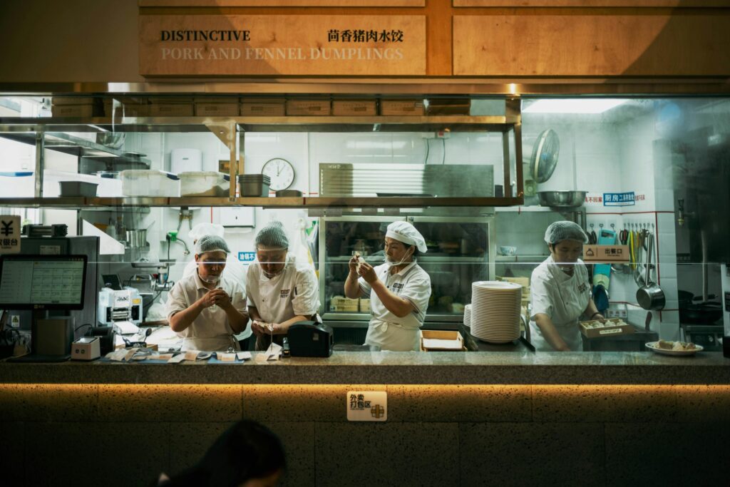 Four chefs wearing white uniforms and hairnets are meticulously preparing dumplings behind a stainless steel kitchen counter. A wooden sign above them identifies their specialty as "Distinctive Pork and Fennel Dumplings".