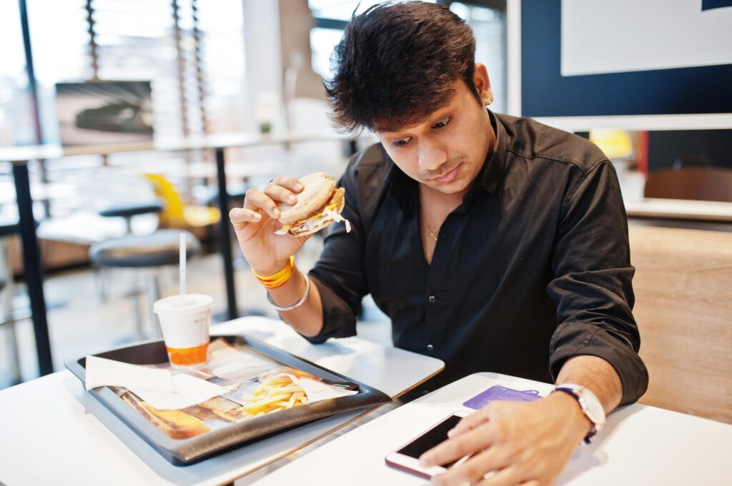 A man in a black shirt sits at a cafe table, looking down at his phone while holding a half-eaten burger. On the table before him is a tray with French fries, napkins, and a cold drink.