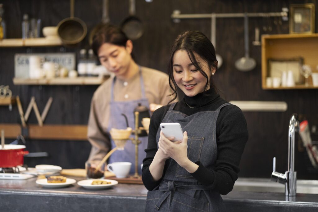 A woman in a dark denim apron smiles while looking at her smartphone in a cozy, rustic kitchen or café setting. In the background, a man in a matching apron is focused on preparing a pour-over coffee near various culinary tools and shelves.