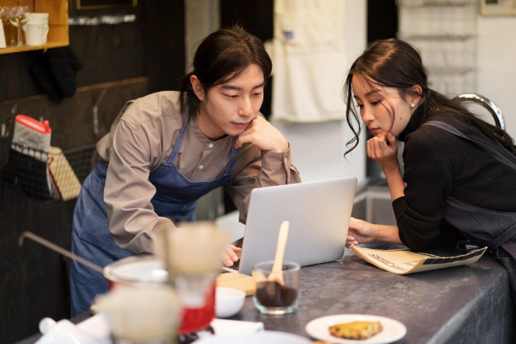 A man and a woman wearing aprons lean over a laptop on a restaurant counter, appearing deeply focused on the screen. The cozy, dimly lit interior features various kitchen tools and a small plate of food in the foreground.
