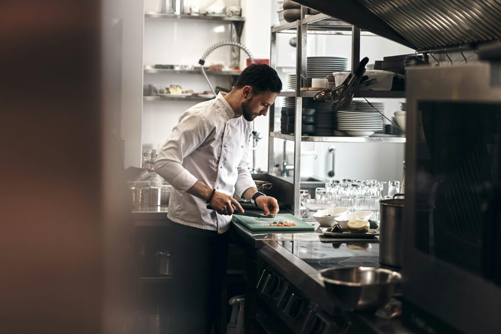 A professional chef in a white uniform is focused on finely dicing ingredients on a green cutting board. He is working in a busy, well-lit commercial kitchen filled with stainless steel equipment, stacked plates, and glassware.
