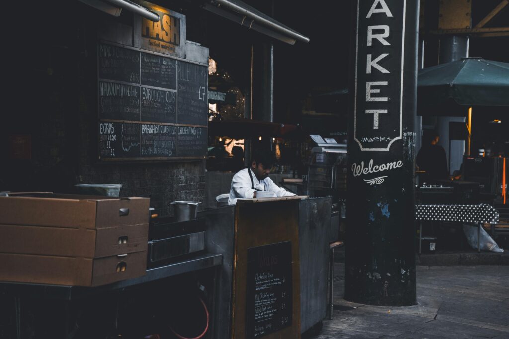 A chef in a white uniform works behind a stall at an urban street food market. Hand-written chalkboard menus and a large pillar marked "MARKET" frame the moody, shadowed scene.