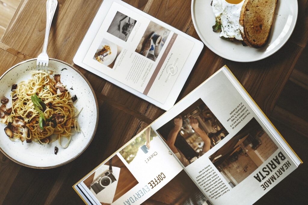 A flat-lay view shows a wooden table set with plates of pasta and eggs alongside an open book and a tablet. The lifestyle composition blends a leisurely meal with reading materials focused on coffee and photography.