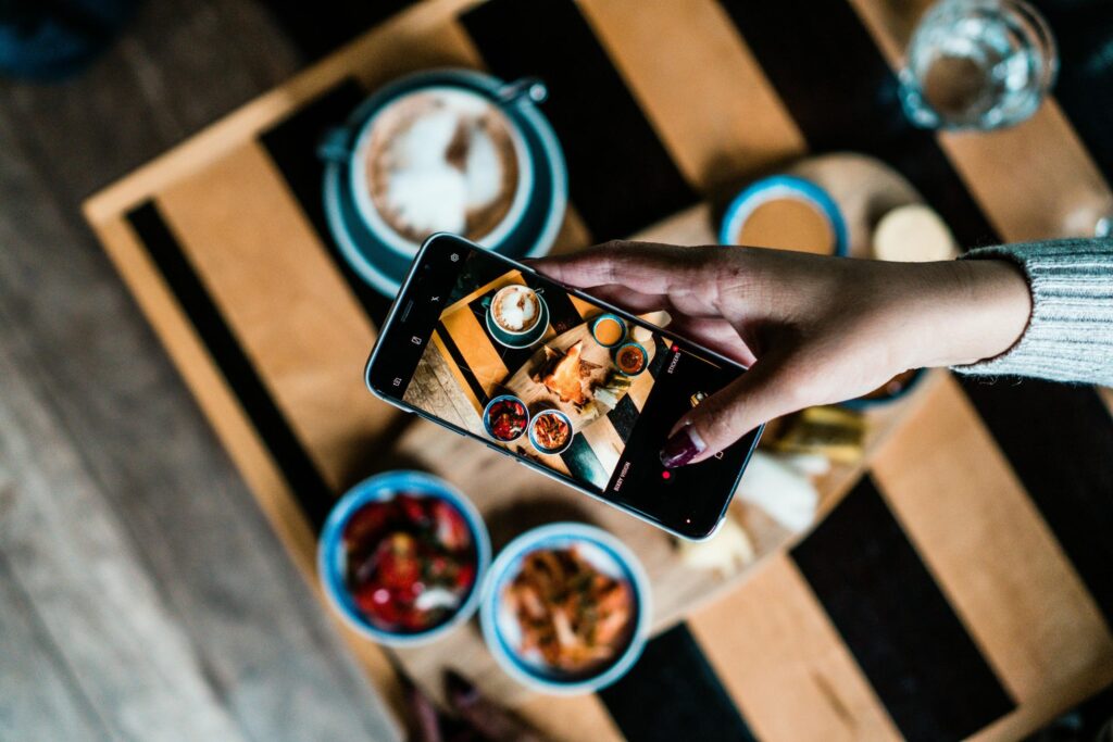 A person's hand holds a smartphone to capture a top-down photo of a diverse breakfast spread arranged on a striped wooden table. The scene features various small bowls of food, coffee with latte art, and toast.
