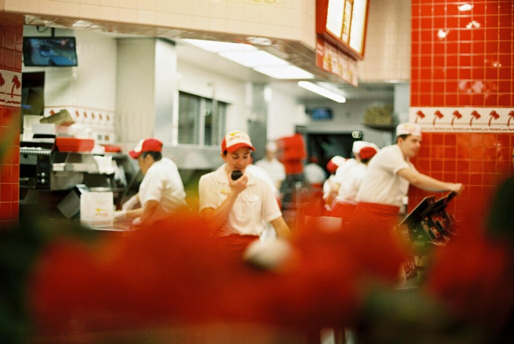 Staff in red and white uniforms work behind the counter of an In-N-Out Burger restaurant. The scene is viewed through a soft, blurry foreground of red flowers.