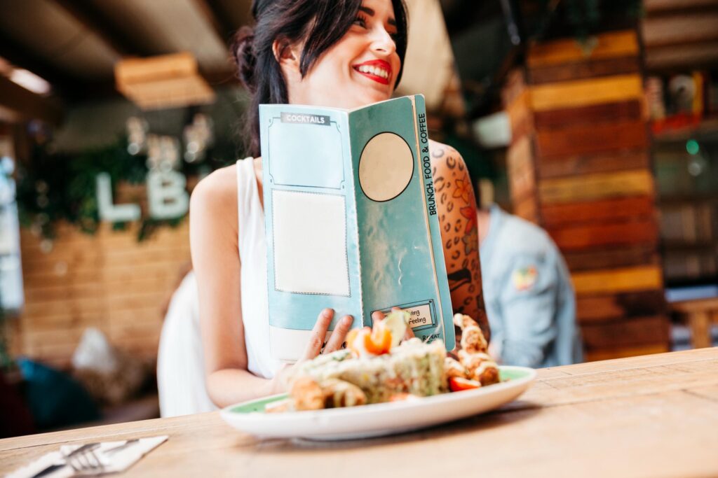 A smiling woman with dark hair and a tattoo on her arm holds a teal menu while seated at a wooden table. A plate of colorful food sits in the foreground within a rustic, warm-toned cafe or restaurant setting.